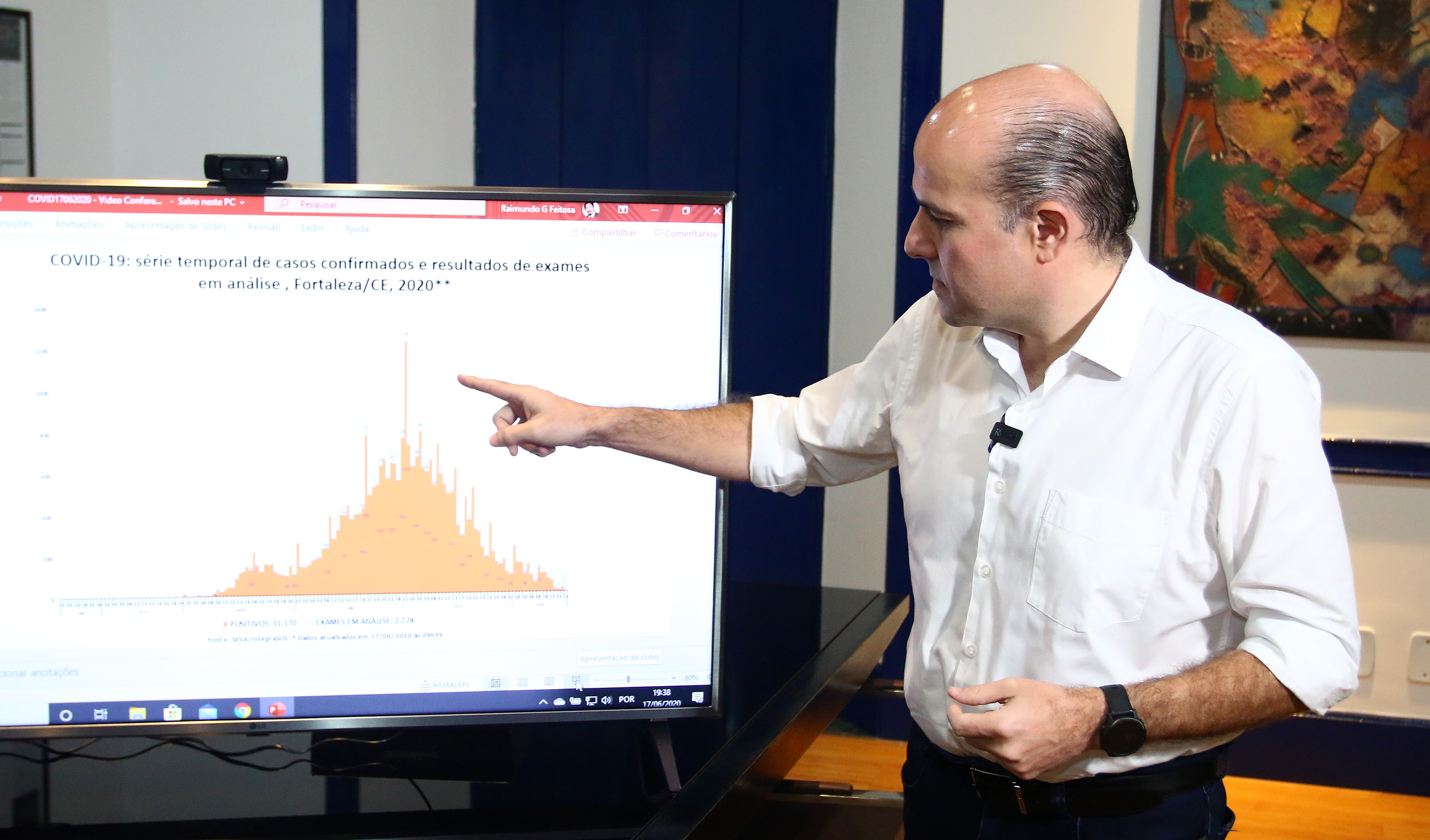 prefeito em pé apontando para televisão que mostra um gráfico